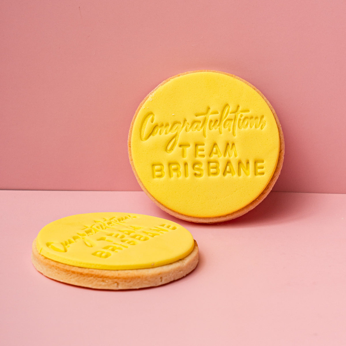 Two yellow cookies with 'Congratulations Team Brisbane' text on a pink background