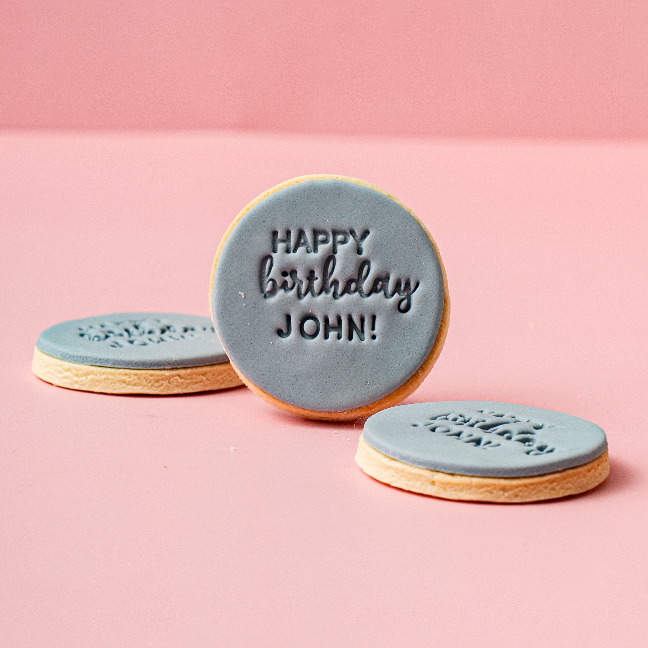 Three round blue cookies with 'Happy Birthday John!' text on a pink background