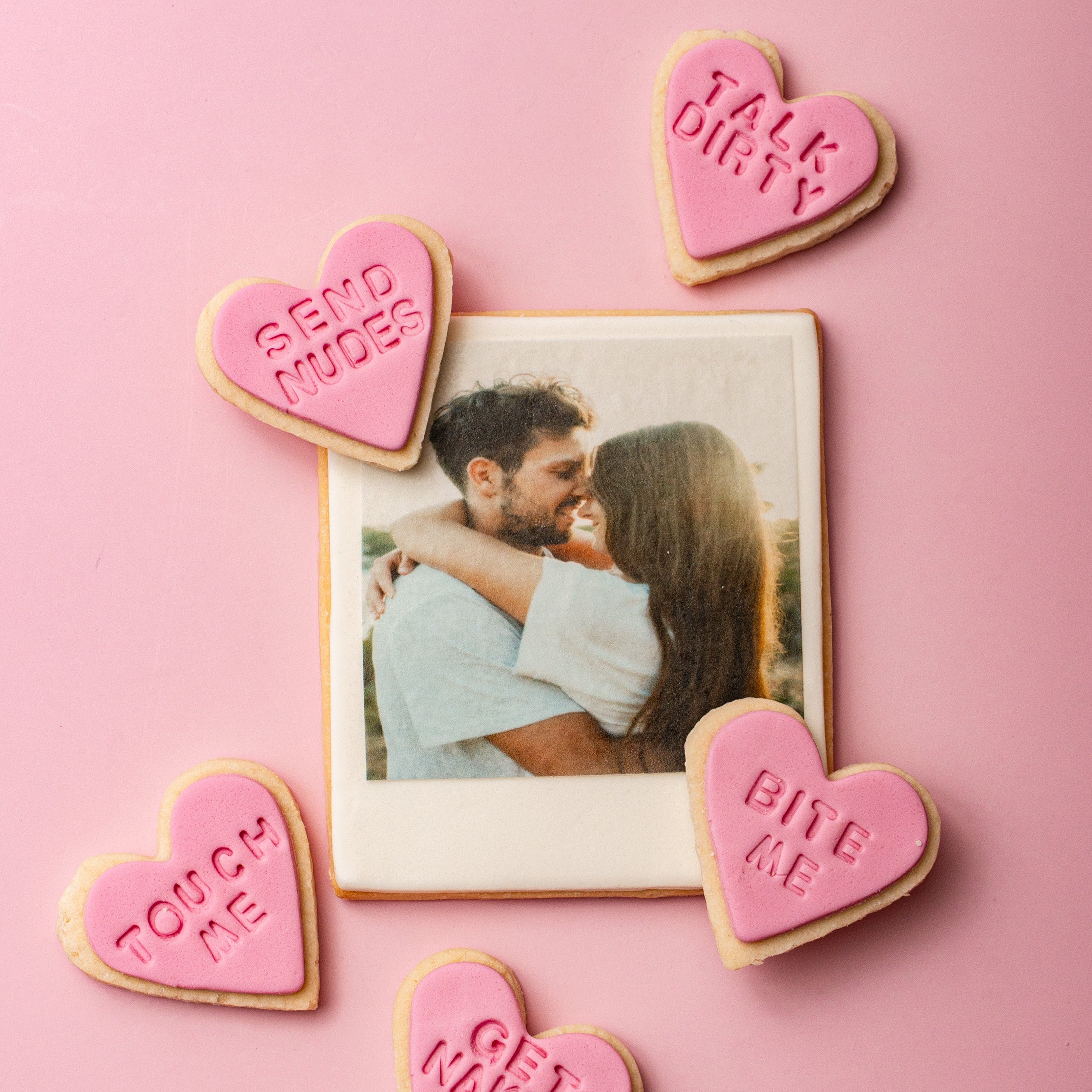 Heart-shaped cookies with suggestive text around a photo of a couple on a pink background