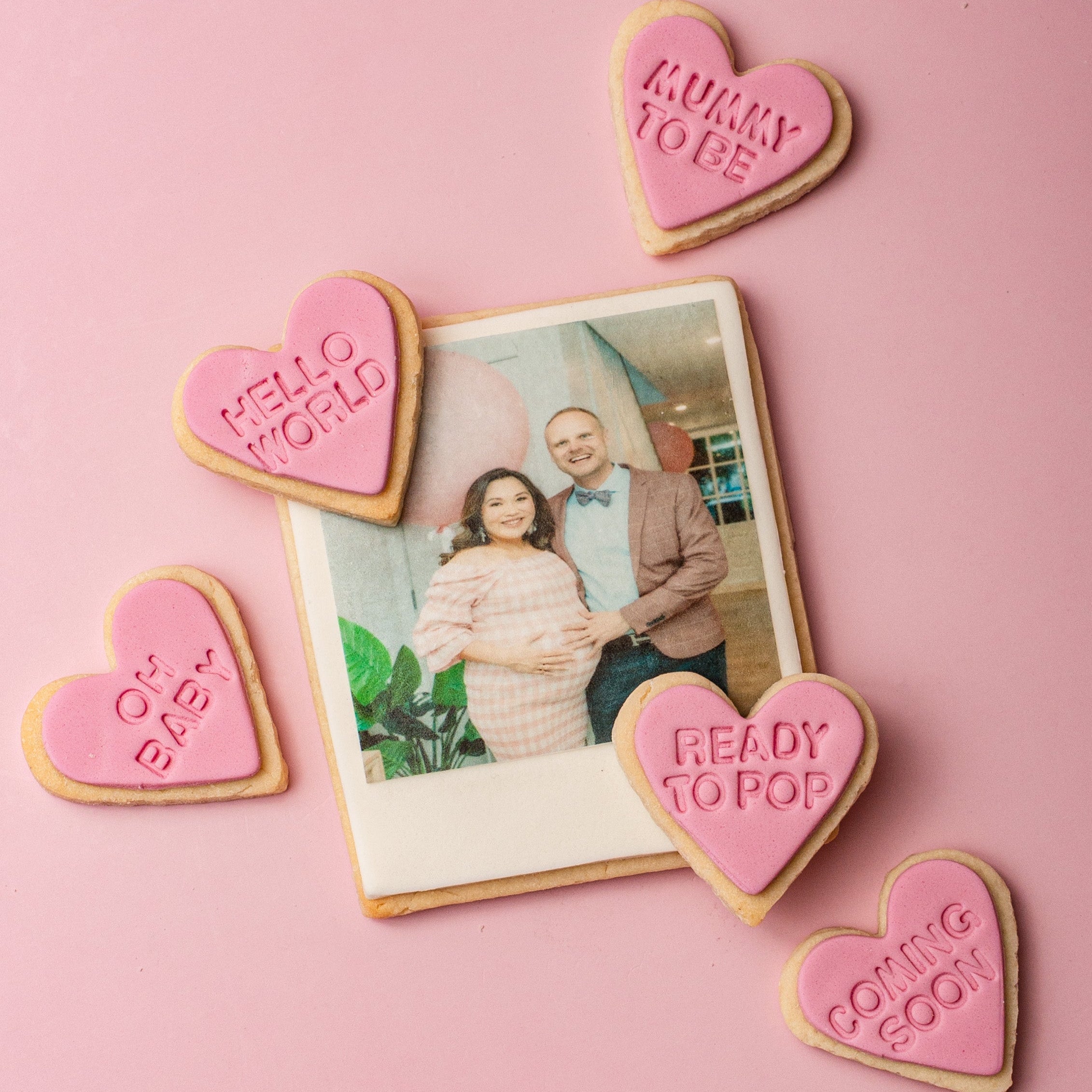 Heart-shaped cookies with pregnancy announcements around a photo of a couple on a pink background