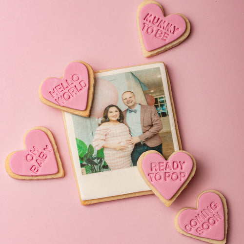 Heart-shaped cookies with pregnancy announcements around a photo of a couple on a pink background