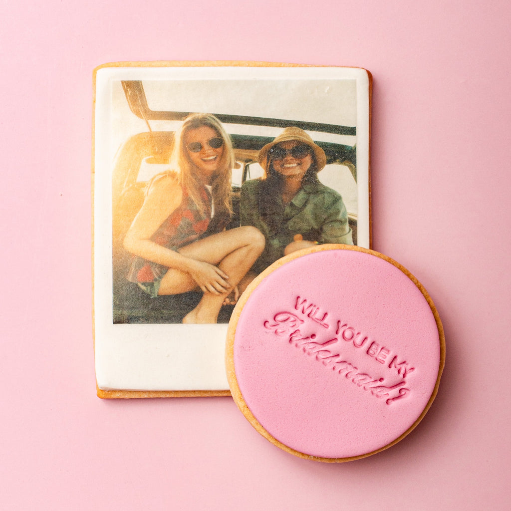 Cookie with 'Will you be my bridesmaid?' text next to a photo of two women on a pink background