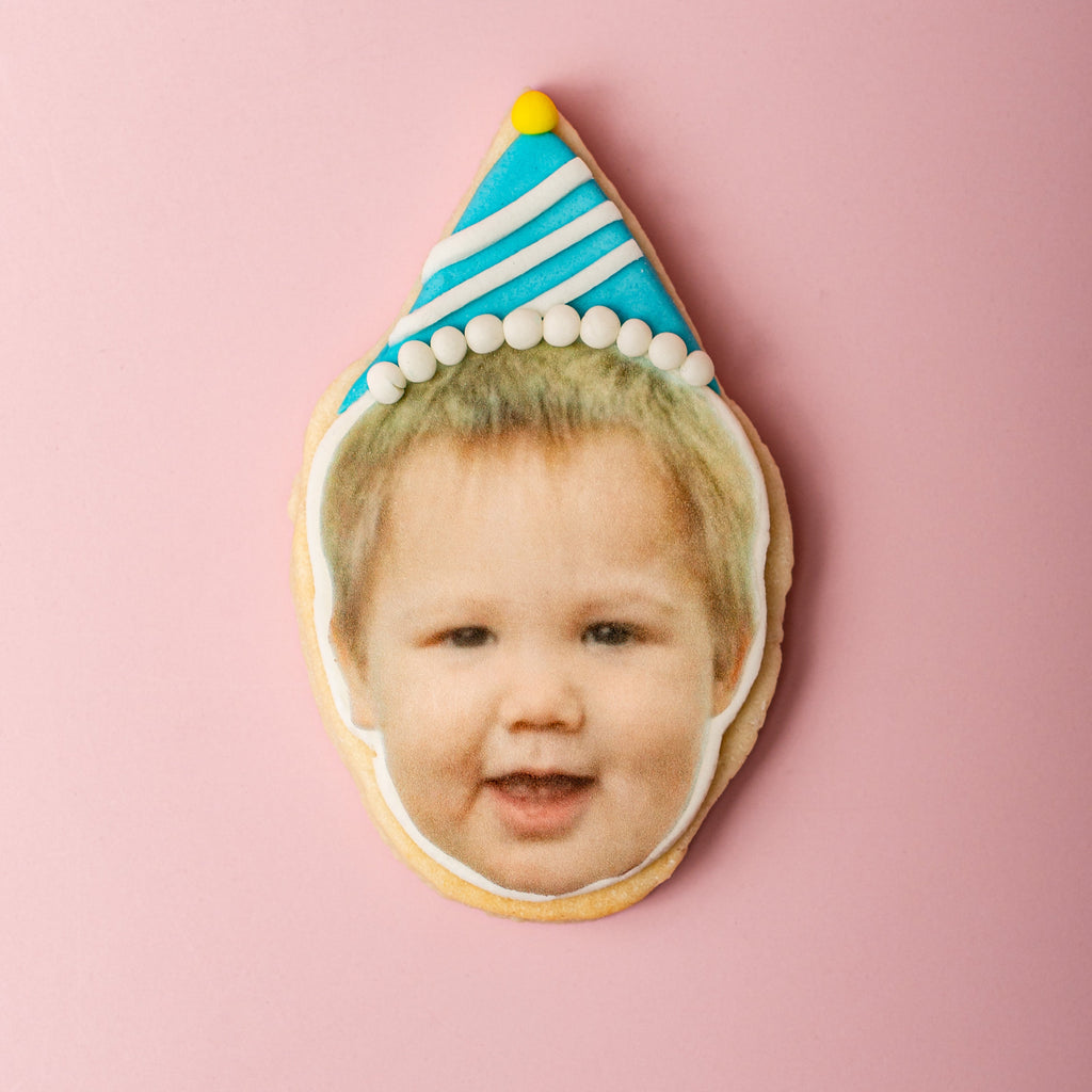Cookie with a face printed on it, featuring a child's face wearing a birthday hat, on a pink background