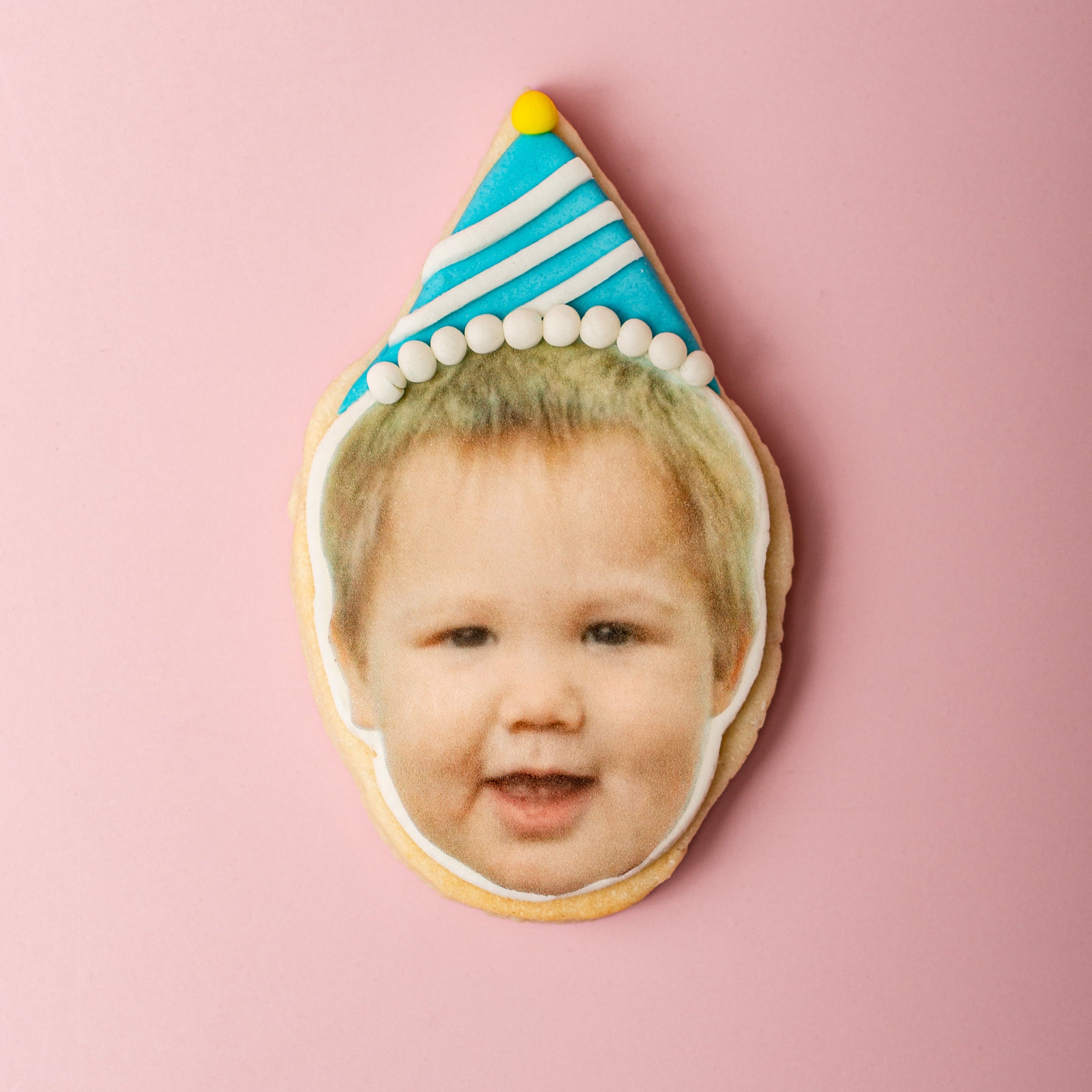 Cookie with a face printed on it, featuring a child's face wearing a birthday hat, on a pink background